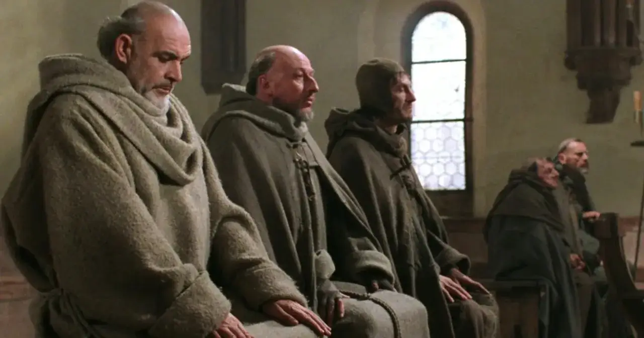 Wilhelm z Baskerville i inni mnisi w szarych habitach siedzą w kościele, pogrążeni w modlitwie.