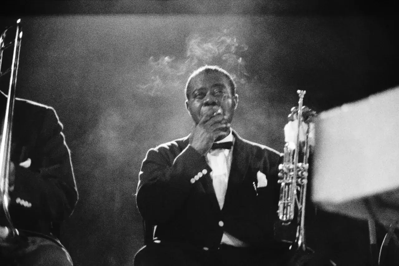 Louis Armstrong, a legendary jazz musician, takes a moment to smoke a cigarette during a performance, his trumpet resting beside him.