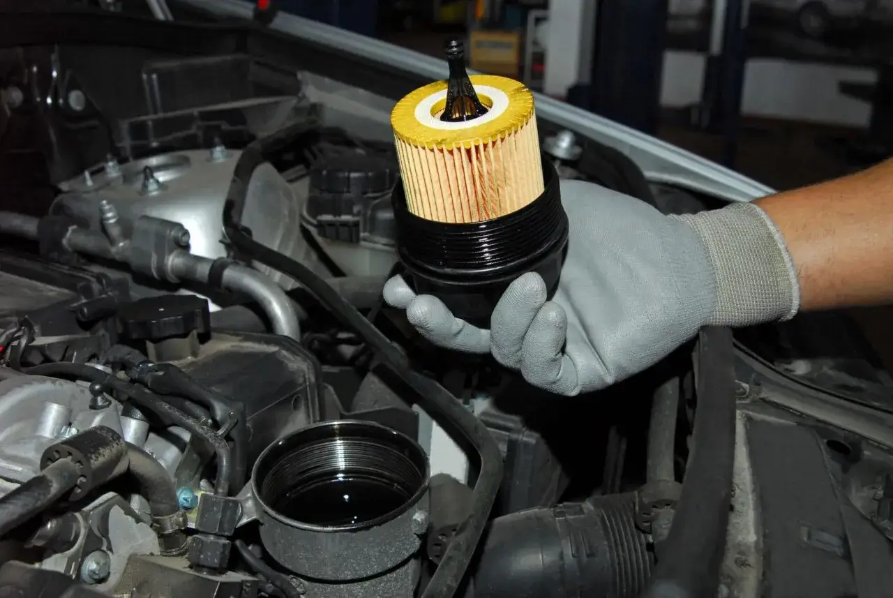 Mano con guanto grigio tiene il filtro olio motore, pronto per la sostituzione. Si vede il vano motore con il contenitore dell'olio.