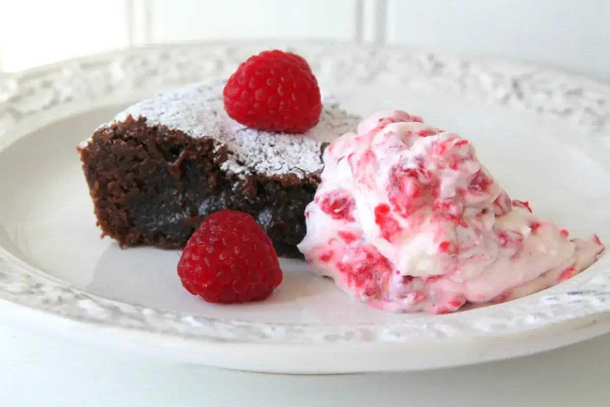 En bit kladdkaka med lakrits, pudrad med florsocker och garnerad med hallon. Serveras med en klick hallongrädde.