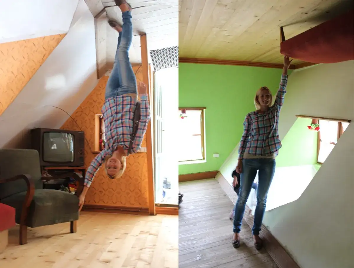 Kobieta w kratowanej koszuli wisi głową w dół w pokoju, który wygląda jak dom do góry nogami. Obok stoi fotel i telewizor.