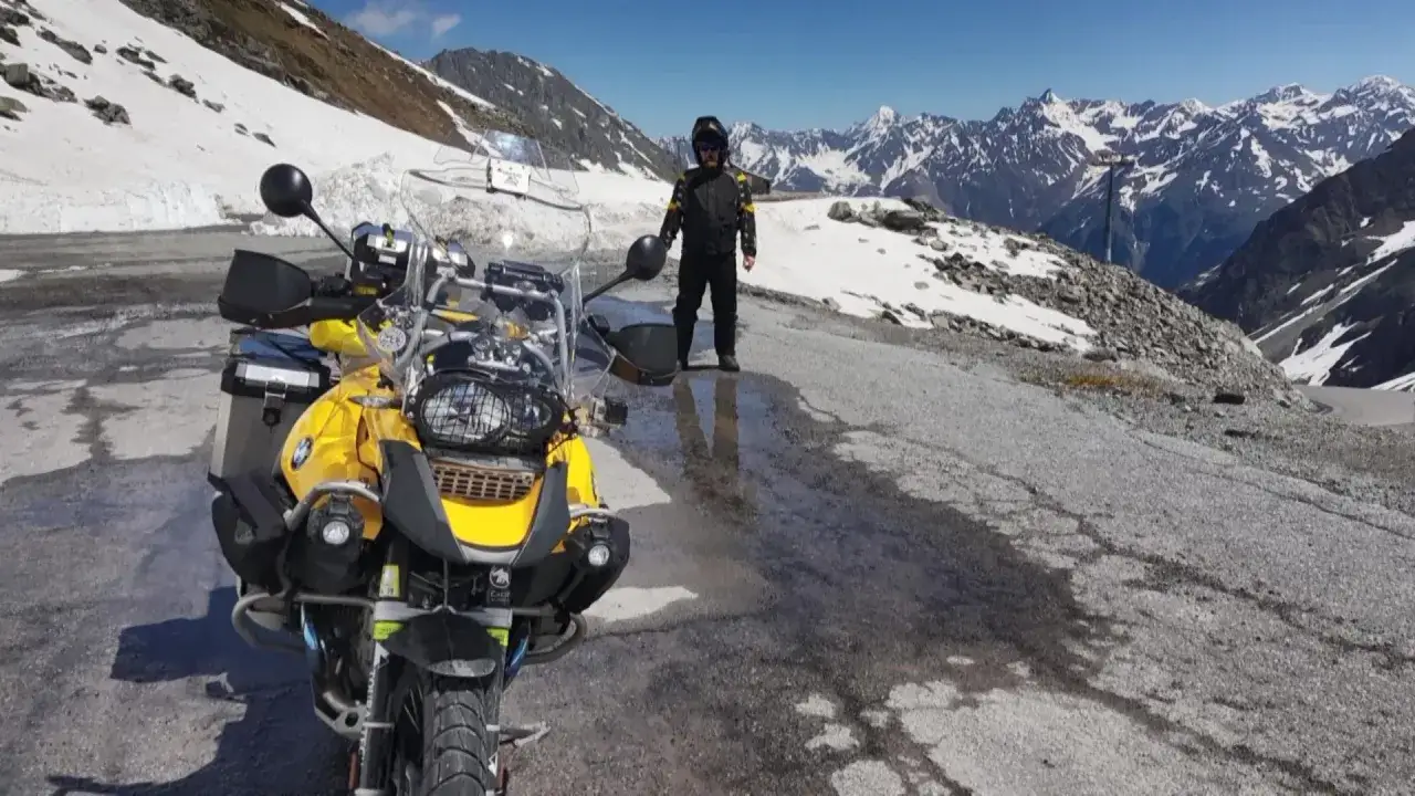 Motociclista in piedi vicino alla sua moto gialla sulla &Ouml;tztaler Gletscherstra&szlig;e, circondato da montagne innevate e un cielo azzurro.