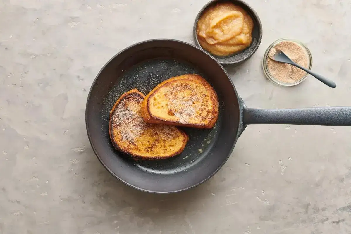 Deux tranches de pain doré dans une poêle, prêtes pour une recette de pain perdu. Compote de pommes et sucre à la cannelle à côté.