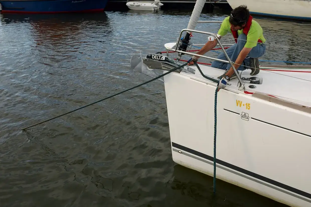 Un homme en gilet de sauvetage ajuste l'amarrage du bateau avec une pendille. L'ancre Delta est visible à l'avant.