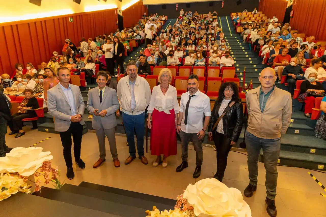Un grupo de personas posa en el escenario del teatro Los Corrales de Buelna ante un p&uacute;blico sentado.