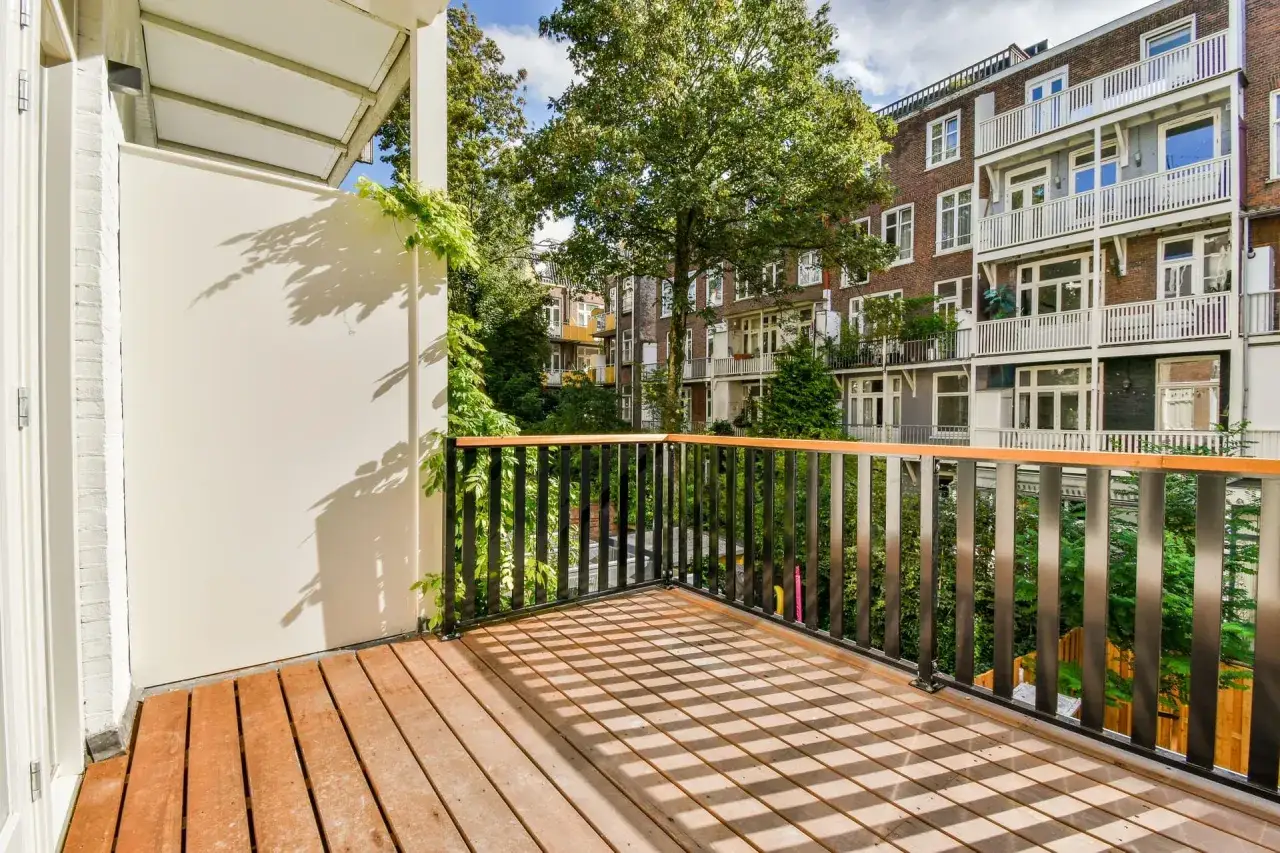 Drewniany balkon z czarną balustradą, z którego widać kamienice i drzewa. Czy balkon jest częścią mieszkania? Tak, to idealne miejsce na poranną kawę.
