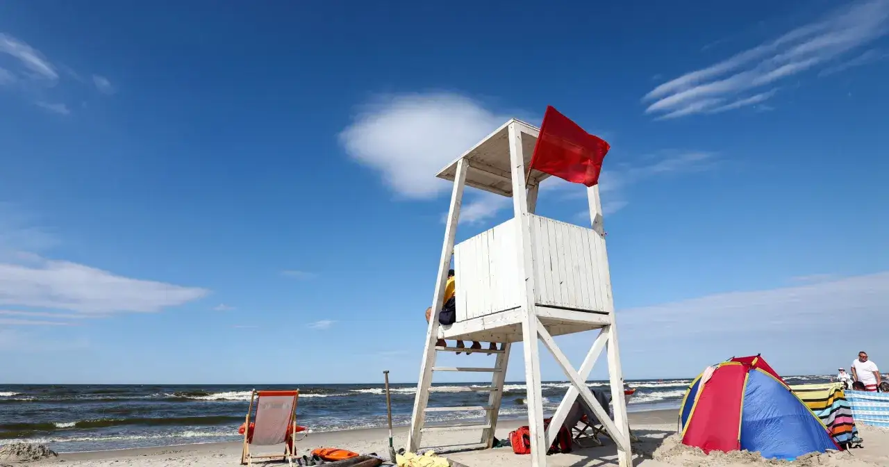 Czerwona flaga nad morzem oznacza zakaz kąpieli. Na plaży biała wieża ratownika, leżaki i namioty.
