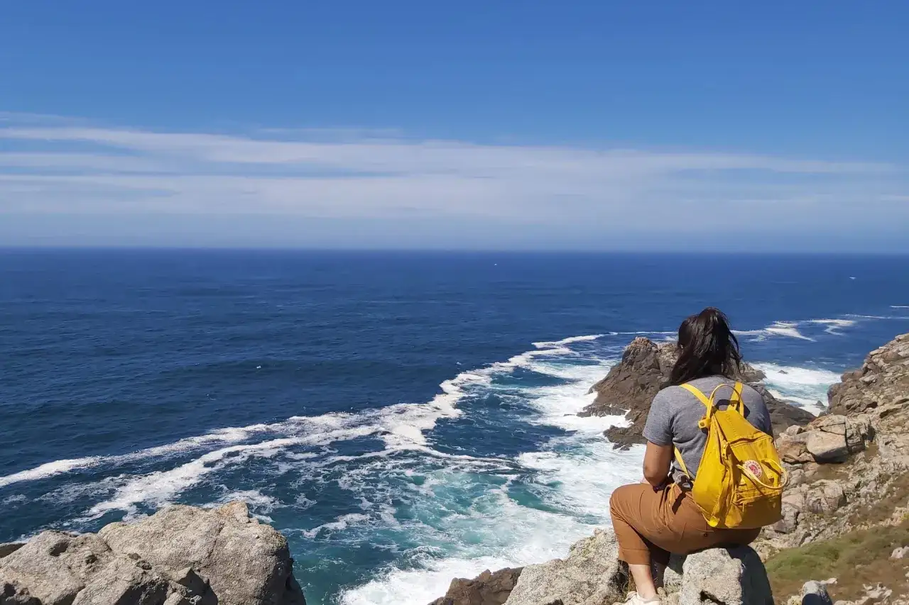 Vue sur l'océan depuis le **Camino dos Faros**. Une randonneuse, sac jaune sur le dos, contemple les vagues déferlant sur la côte rocheuse.
