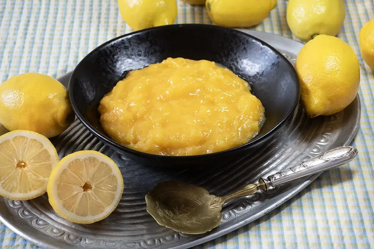 Un tazón negro lleno de cremoso lemon curd, rodeado de limones frescos y cortados. Una cuchara antigua descansa a su lado.