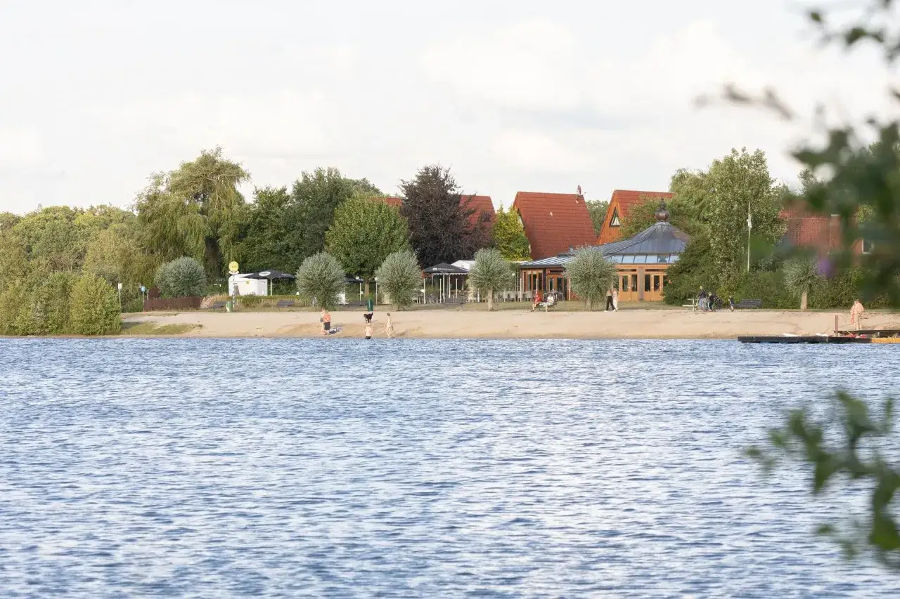 Ein sonniger Tag am See in Gronau. Menschen genie&szlig;en den Strand und das Wasser, w&auml;hrend im Hintergrund Geb&auml;ude und B&auml;ume zu sehen sind.