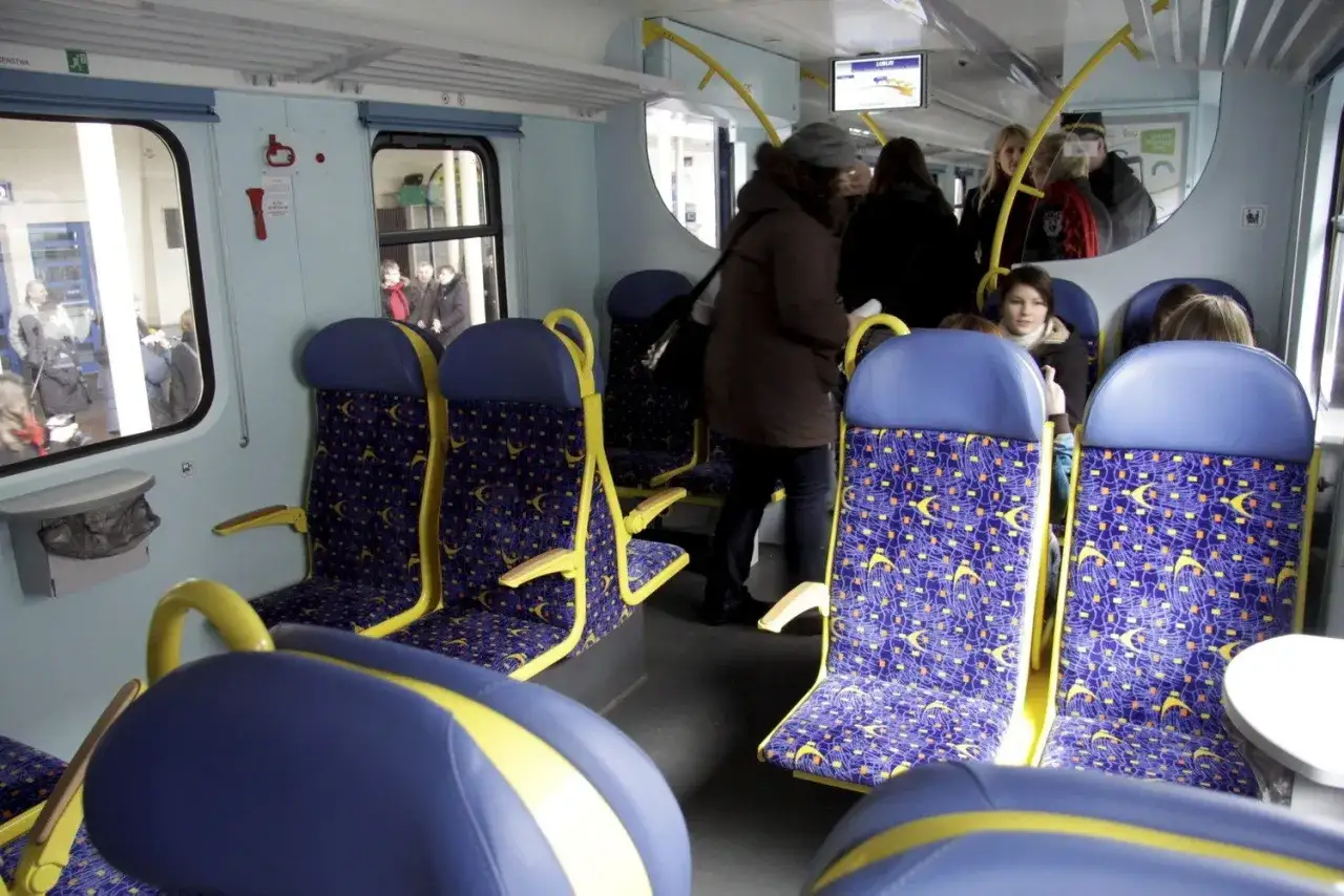 Wnętrze szynobusu z niebieskimi siedzeniami w żółte wzory. Pasażerowie stoją i siedzą, czekając na podróż.