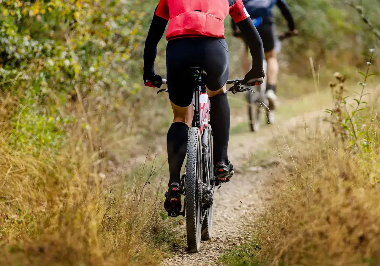 Kolarze w akcji podczas wyścigu gravelowego. Zbliżenie na tylną część roweru i nogi zawodnika w czerwonej koszulce, jadącego po szutrowej ścieżce wśród traw.
