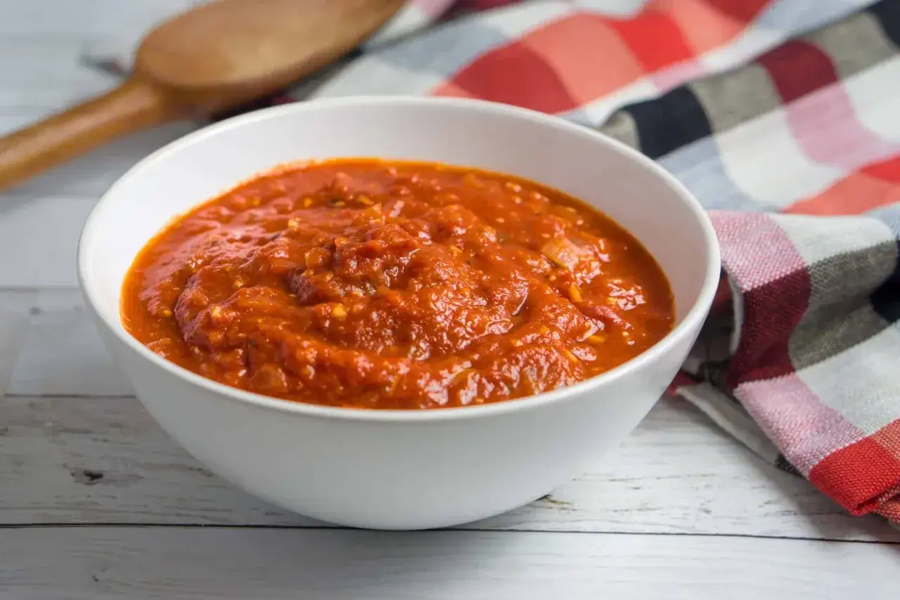 Schüssel mit leckerer San Marzano Pizza Sauce, bereit für die nächste Pizza. Daneben ein Holzlöffel und ein kariertes Tuch.