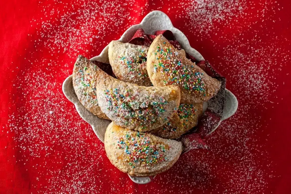 Biscotti tipici calabresi cosparsi di zucchero a velo e confettini colorati, serviti in una ciotola su un telo rosso.