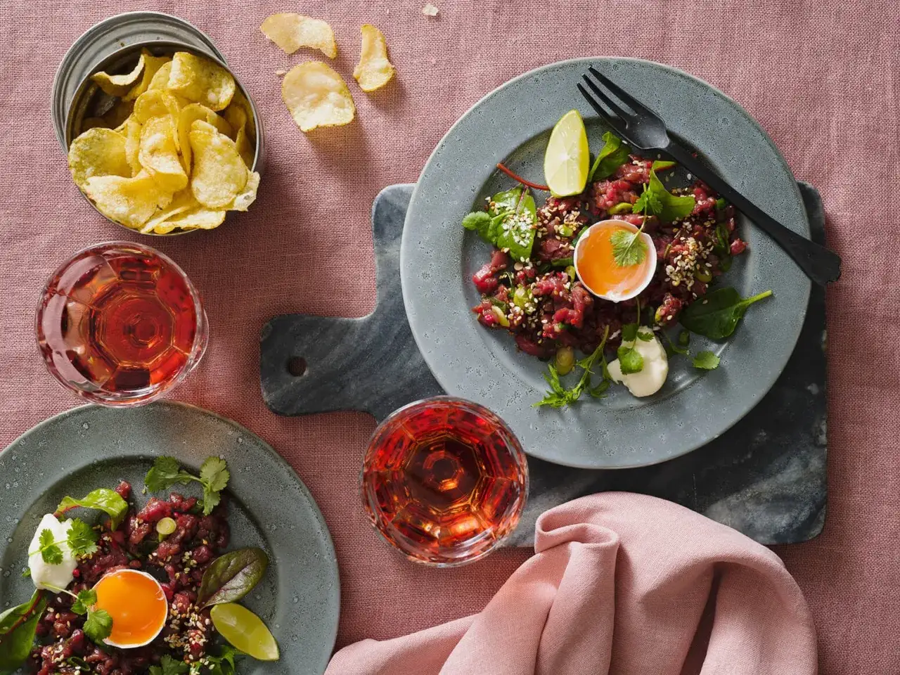Tv&aring; tallrikar med asiatisk r&aring;biff, toppad med &auml;ggula och sesamfr&ouml;n, serveras med ros&eacute;vinsglas och potatischips.