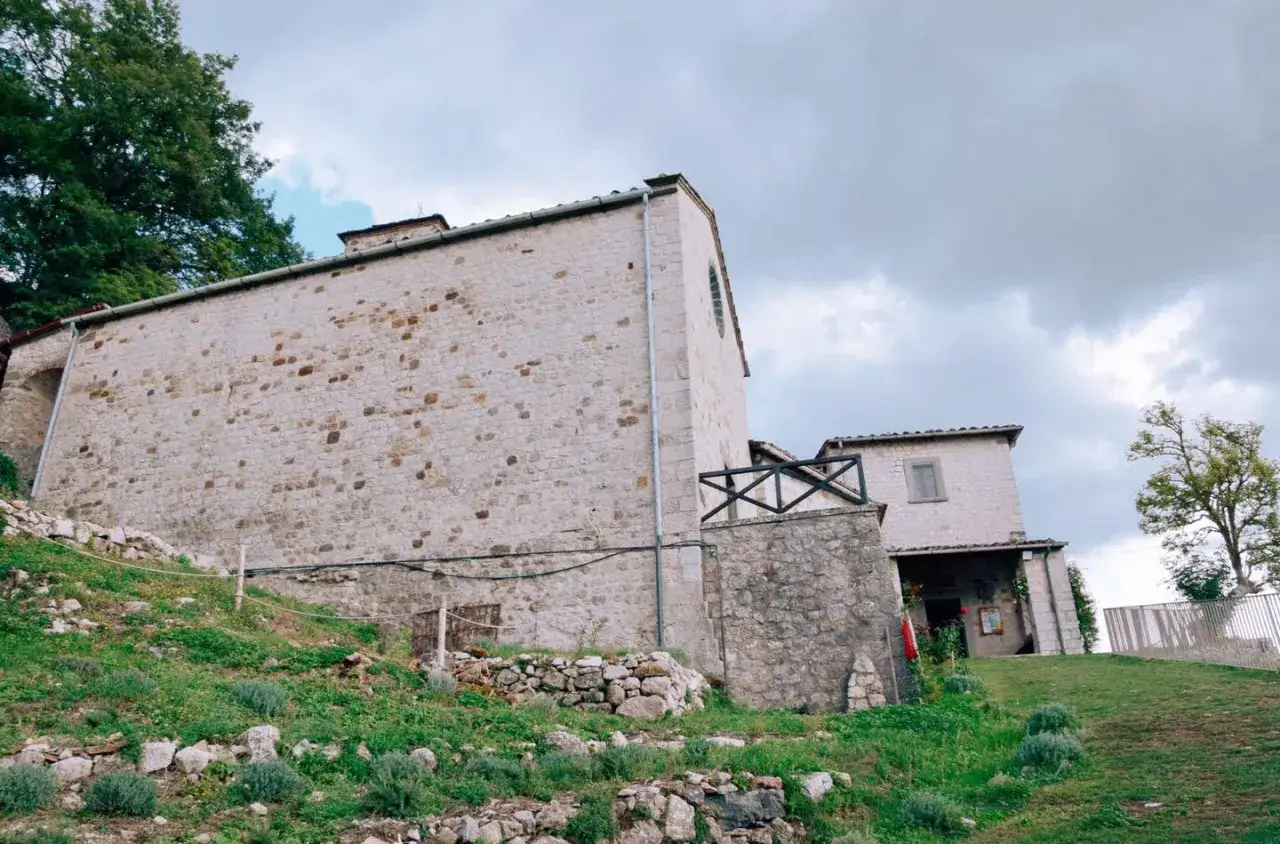 Edifici in pietra con tetto spiovente e un piccolo balcone in legno. Il **eremo di Cerbaiolo** si erge su un pendio erboso, con alberi sullo sfondo.