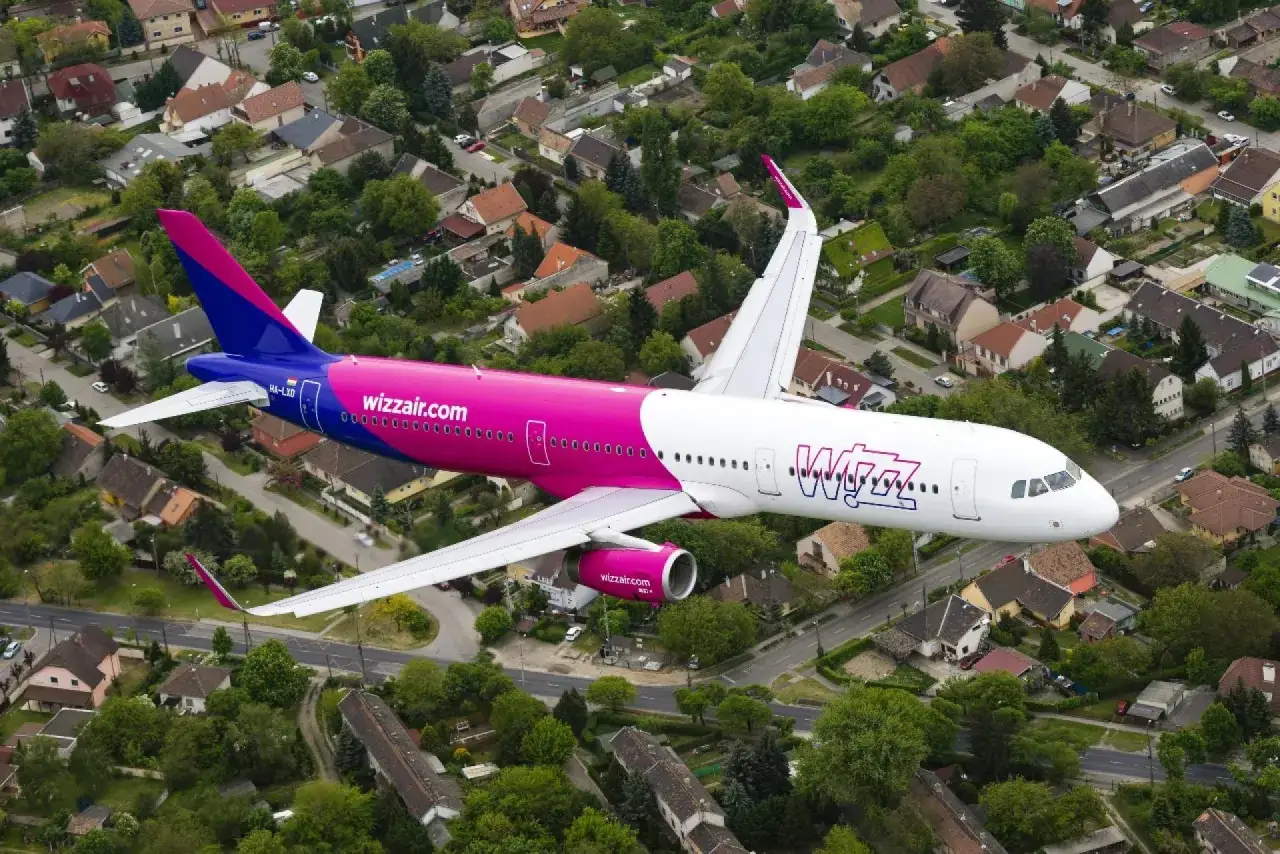 Samoloty Wizzair w locie nad osiedlem domków jednorodzinnych. Różowo-fioletowy kadłub i białe skrzydła.