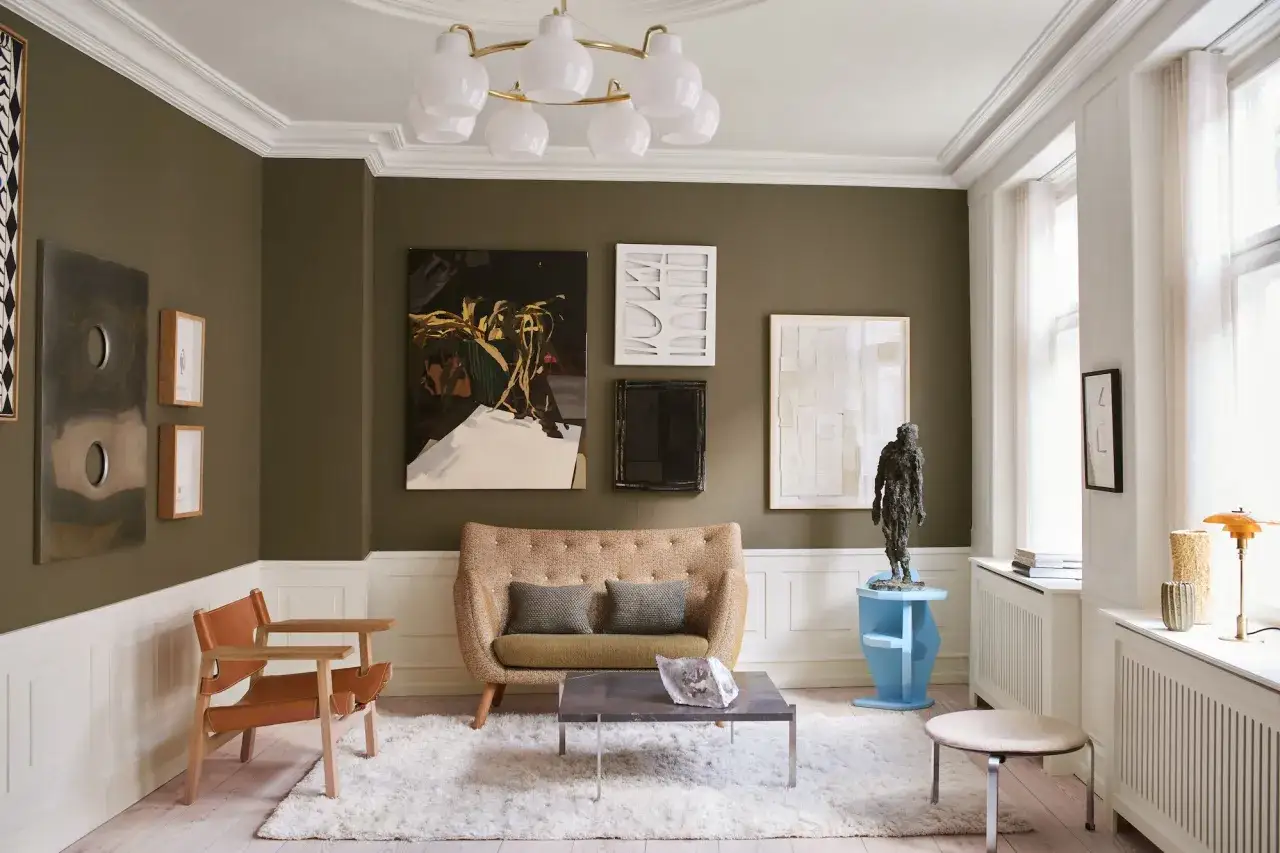 Gemütliches Wohnzimmer mit Kunstwerken, einem beigen Sofa und einem Ledersessel. Warme Farben dominieren die Szene, die durch die Beleuchtung noch verstärkt werden.
