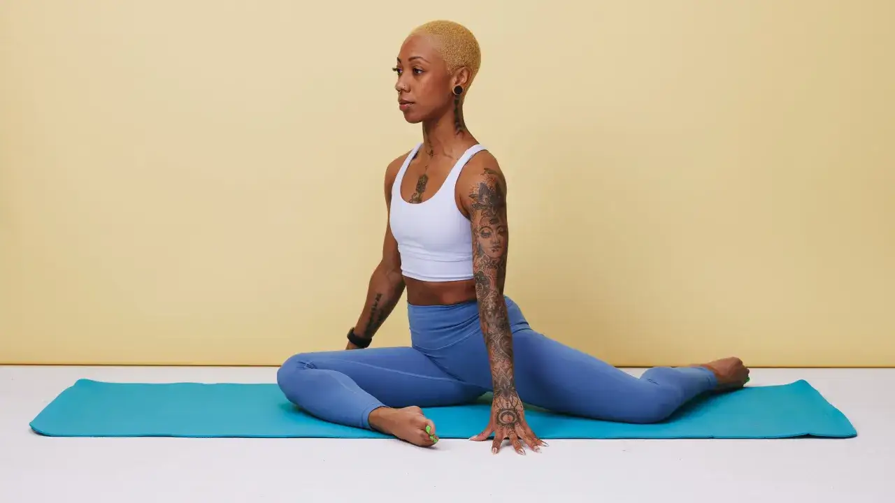 Femme aux tatouages pratiquant un étirement abducteur sur un tapis de yoga bleu.