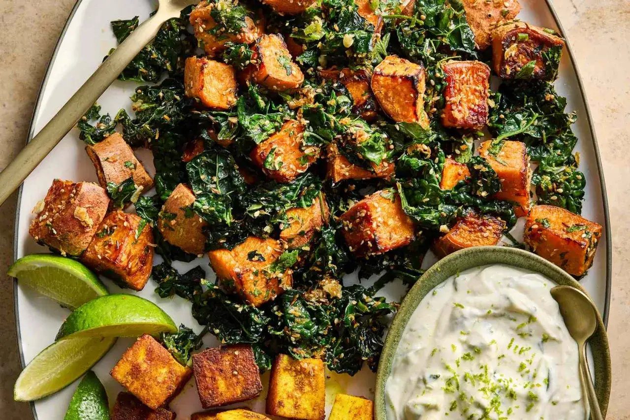 En härlig tallrik med vegetarisk mat: rostade sötpotatisbitar och krispig grönkål, toppad med sesamfrön och serverad med en krämig dipp och limeklyftor.