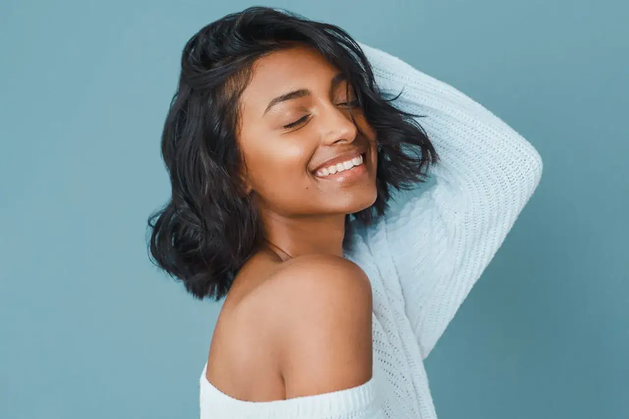 Mujer sonríe con ojos cerrados, luciendo un corte pelo ondulado y un suéter azul claro.