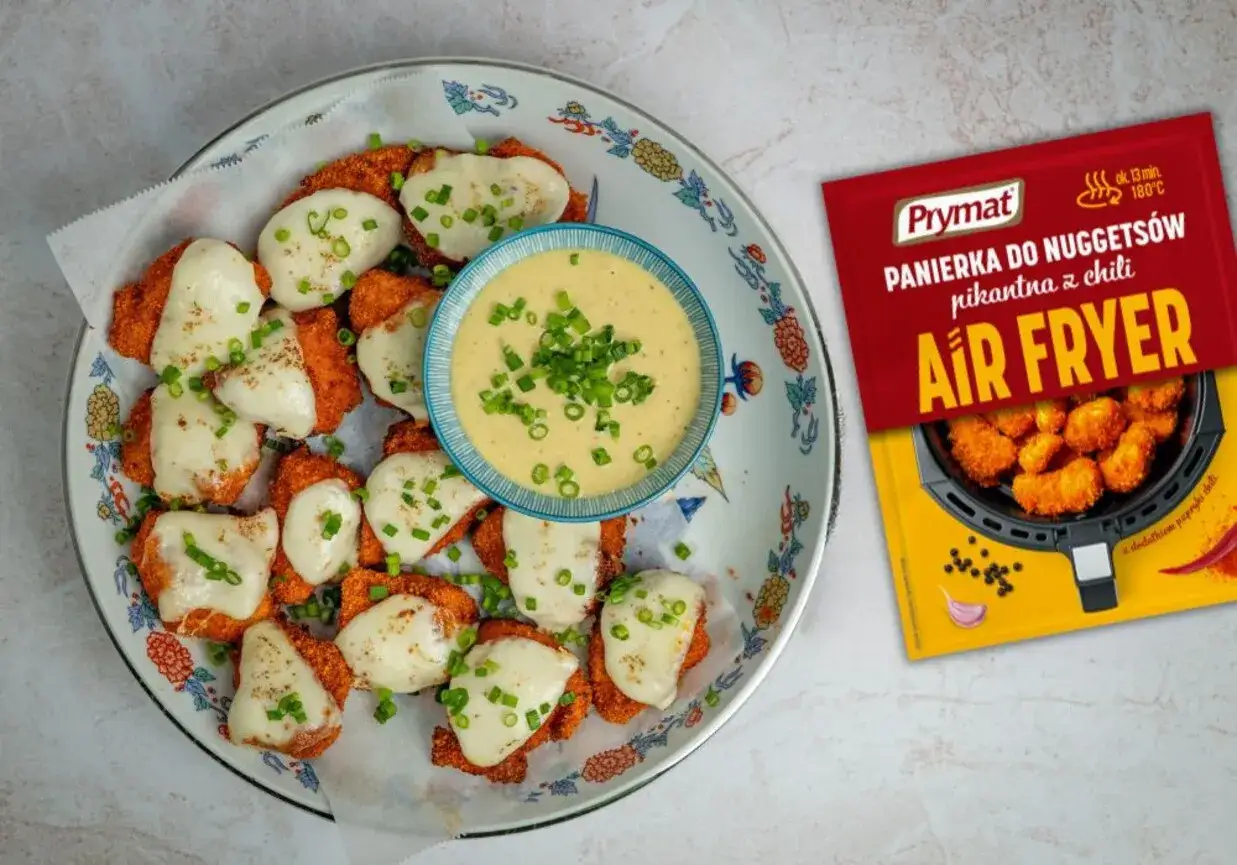 Nuggetsy z serem i szczypiorkiem, obok miseczka z kremowym sosem. Obok opakowanie panierki Prymat do nuggetsów pikantna z chili.
