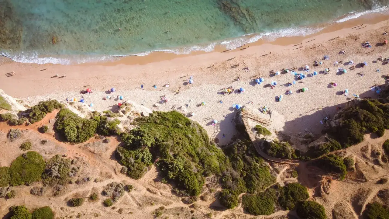 Vista a&eacute;rea de una playa con sombrillas y gente disfrutando del sol. Un camino de madera desciende por la costa, ideal para los mejores camping Andaluc&iacute;a.