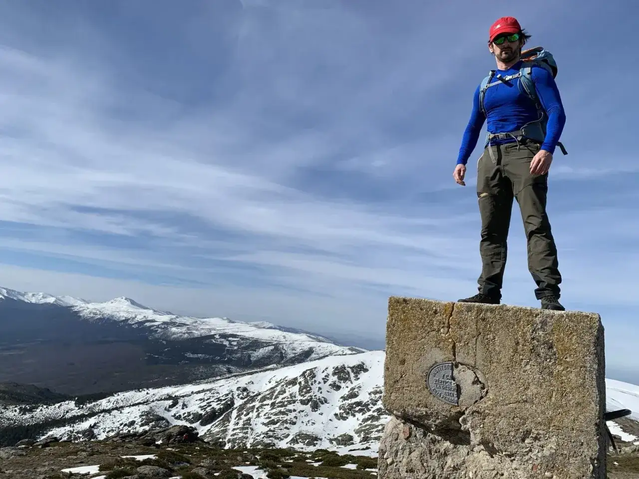 Hombre con mochila en la cima de un pico del nevero, con montañas nevadas de fondo.
