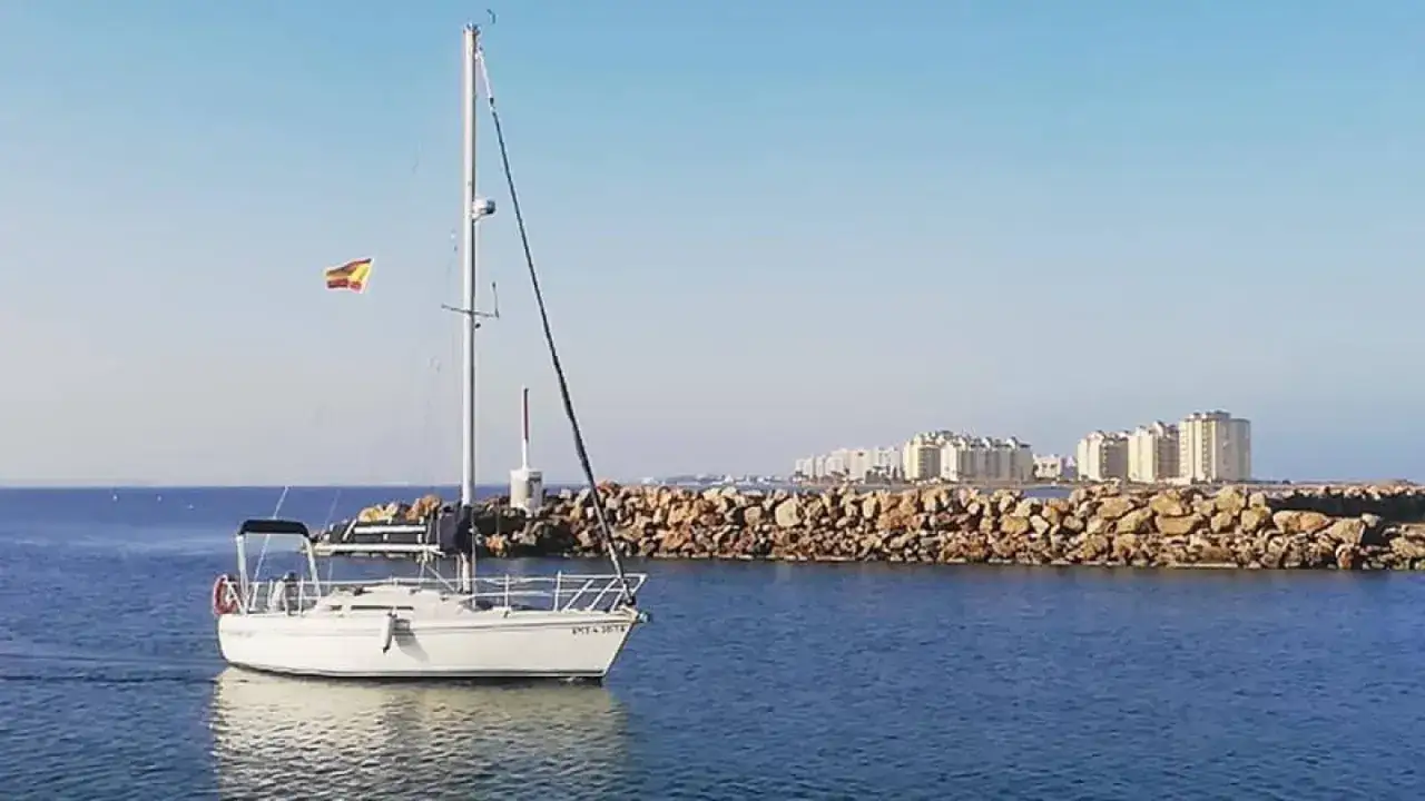 Un voilier Aquila navigue paisiblement en mer, pavillon espagnol au mât, face à des immeubles modernes derrière une digue rocheuse.