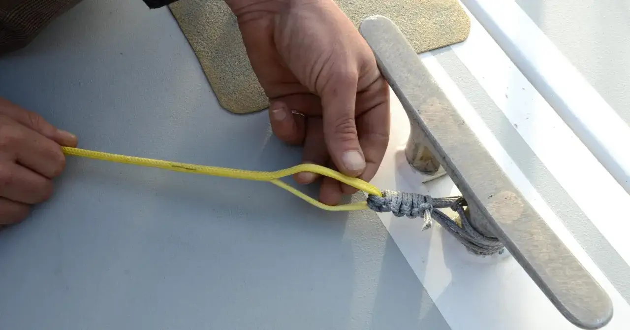 Main d'un marin préparant la ligne de vie d'un voilier, nouant une cordelette jaune à une amarre grise sur un taquet.