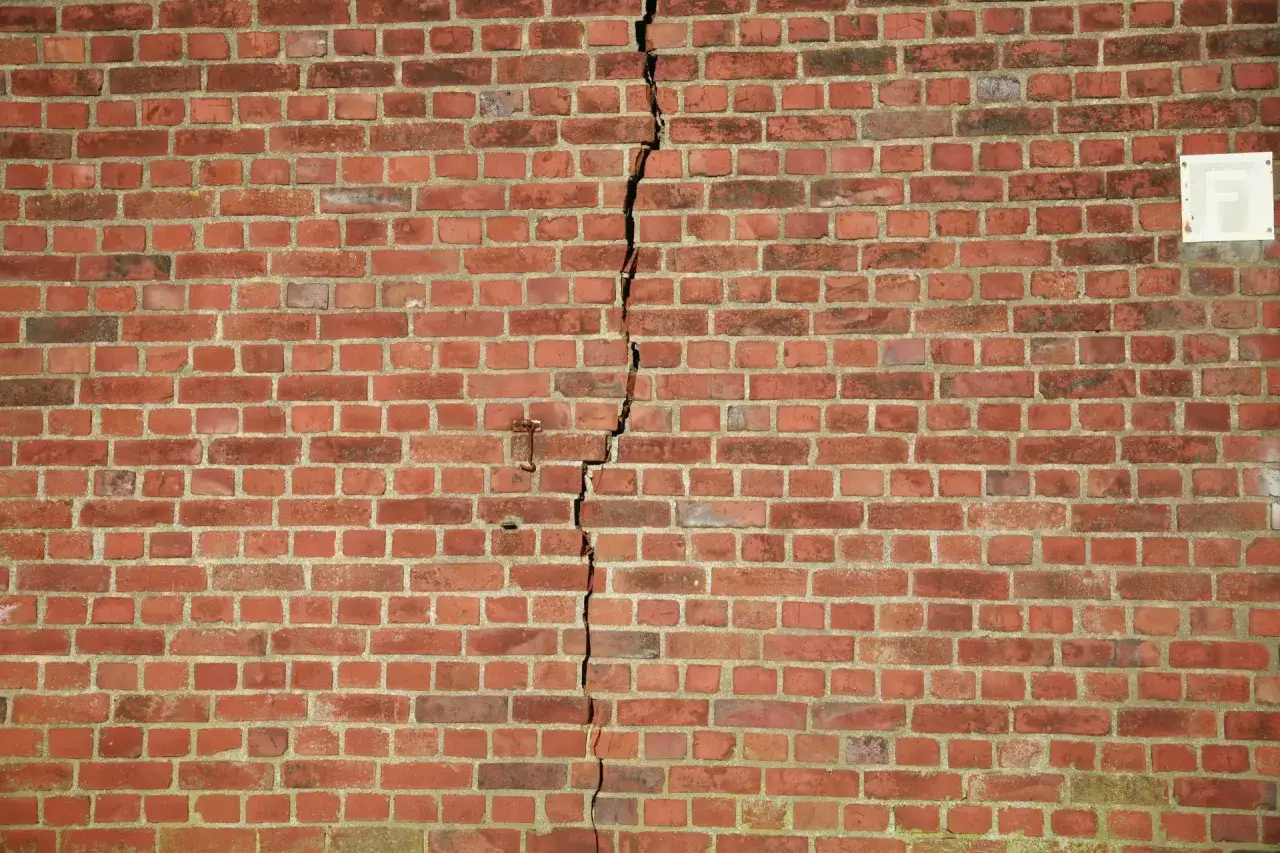 Eine rote Ziegelwand mit deutlichen, vertikalen Rissen. Ein kleines Metallteil ragt aus der Wand nahe der Risse.