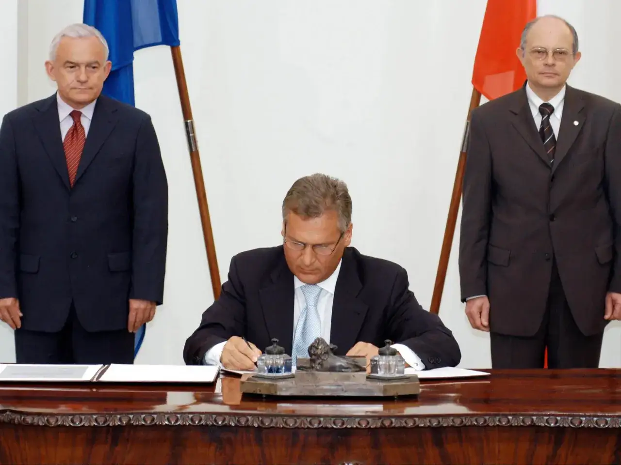 Uroczyste podpisanie dokumentów, gdy Polska weszła do Unii. Trzech mężczyzn w garniturach, obok flagi UE i Polski.