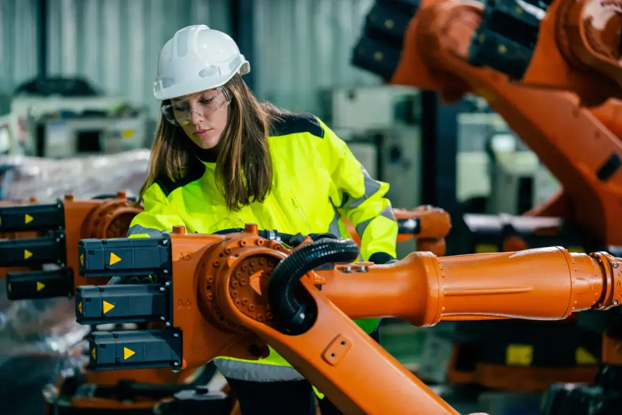 Kobieta w kasku i kamizelce odblaskowej, jako społeczny inspektor pracy, sprawdza stan techniczny ramienia robota przemysłowego.