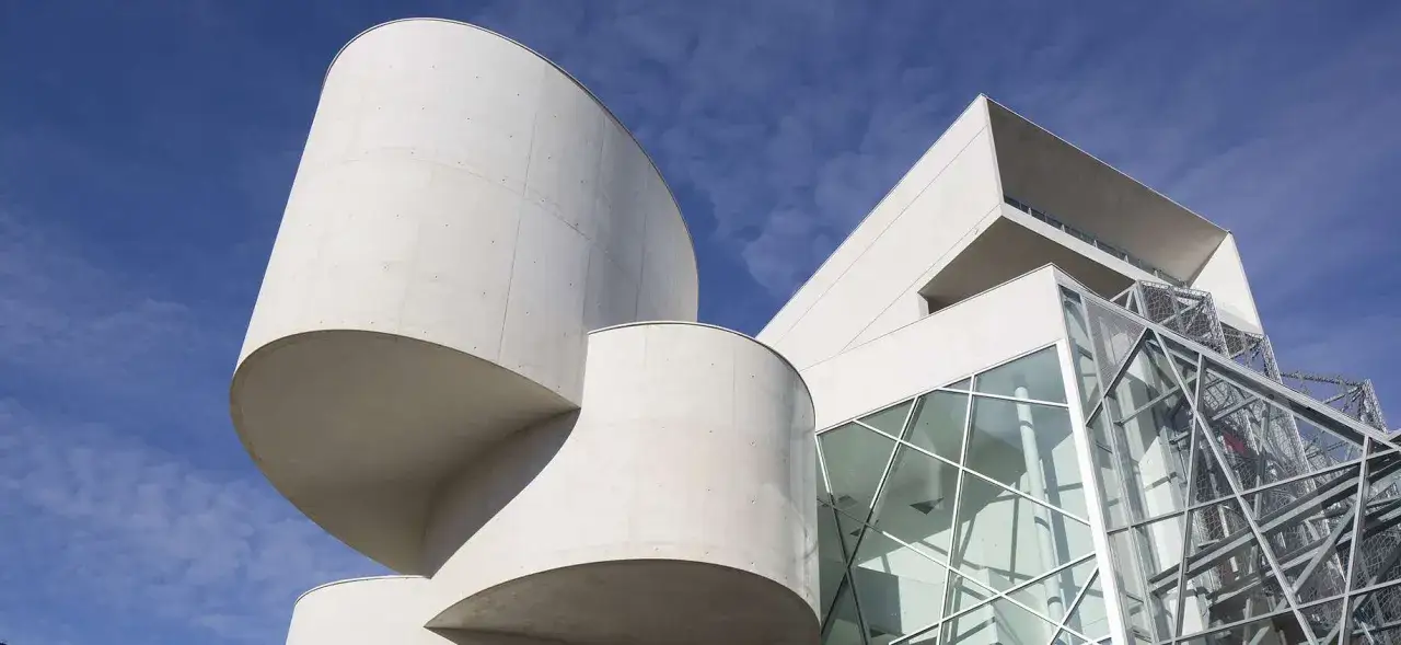 Un bâtiment aux formes audacieuses en béton blanc et verre, capturé par un photographe architecture. Le ciel bleu et nuageux contraste avec les lignes modernes.