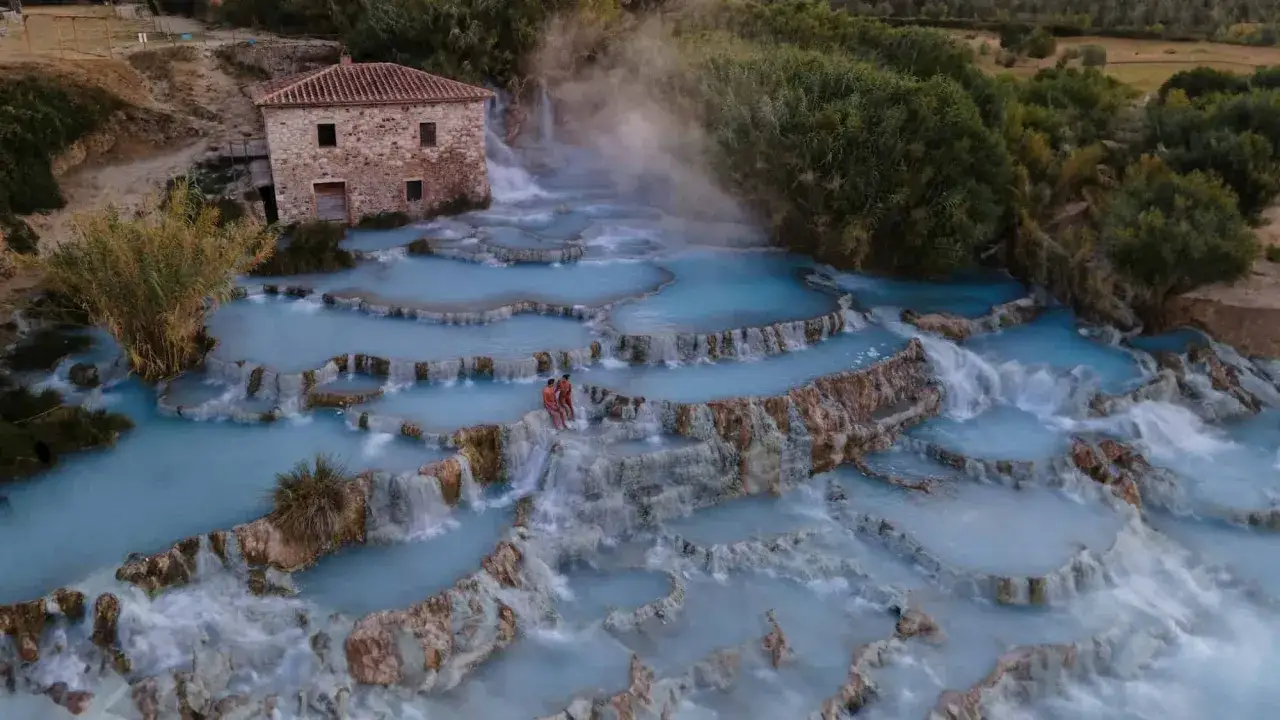Cascate naturali con acqua azzurra e vapore, un casolare in pietra e due persone sedute sulle rocce alle terme di Saturnia.