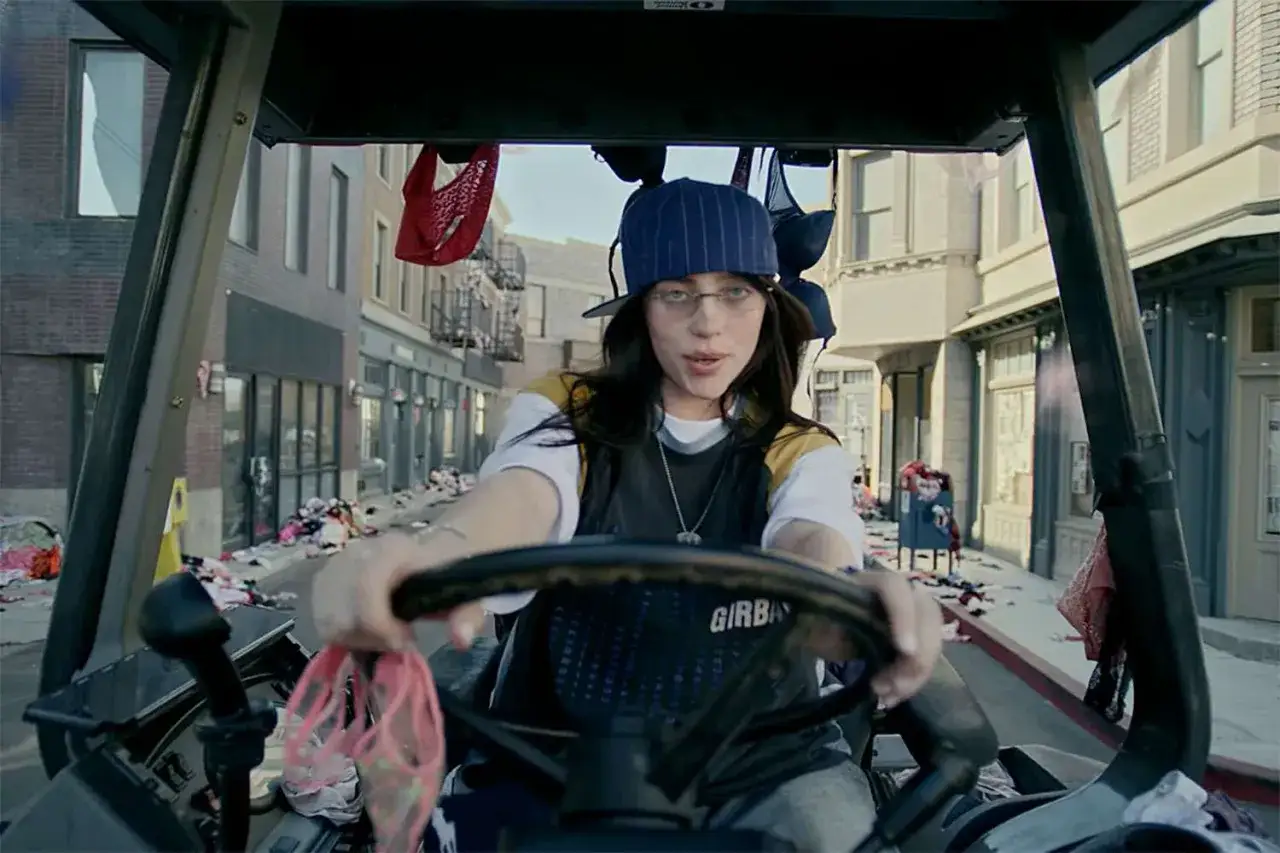 Billie Eilish, jak brat, prowadzi wózek widłowy przez ulicę zasypaną ubraniami.