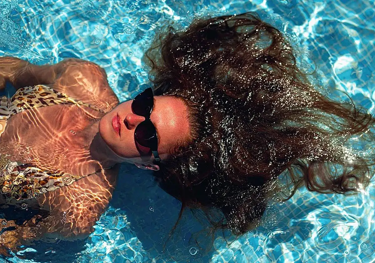 Mujer flotando en piscina, con gafas de sol y bikini. Su pelo oscuro se esparce, brillando bajo el sol, como si sufriera de caida de pelo en verano.