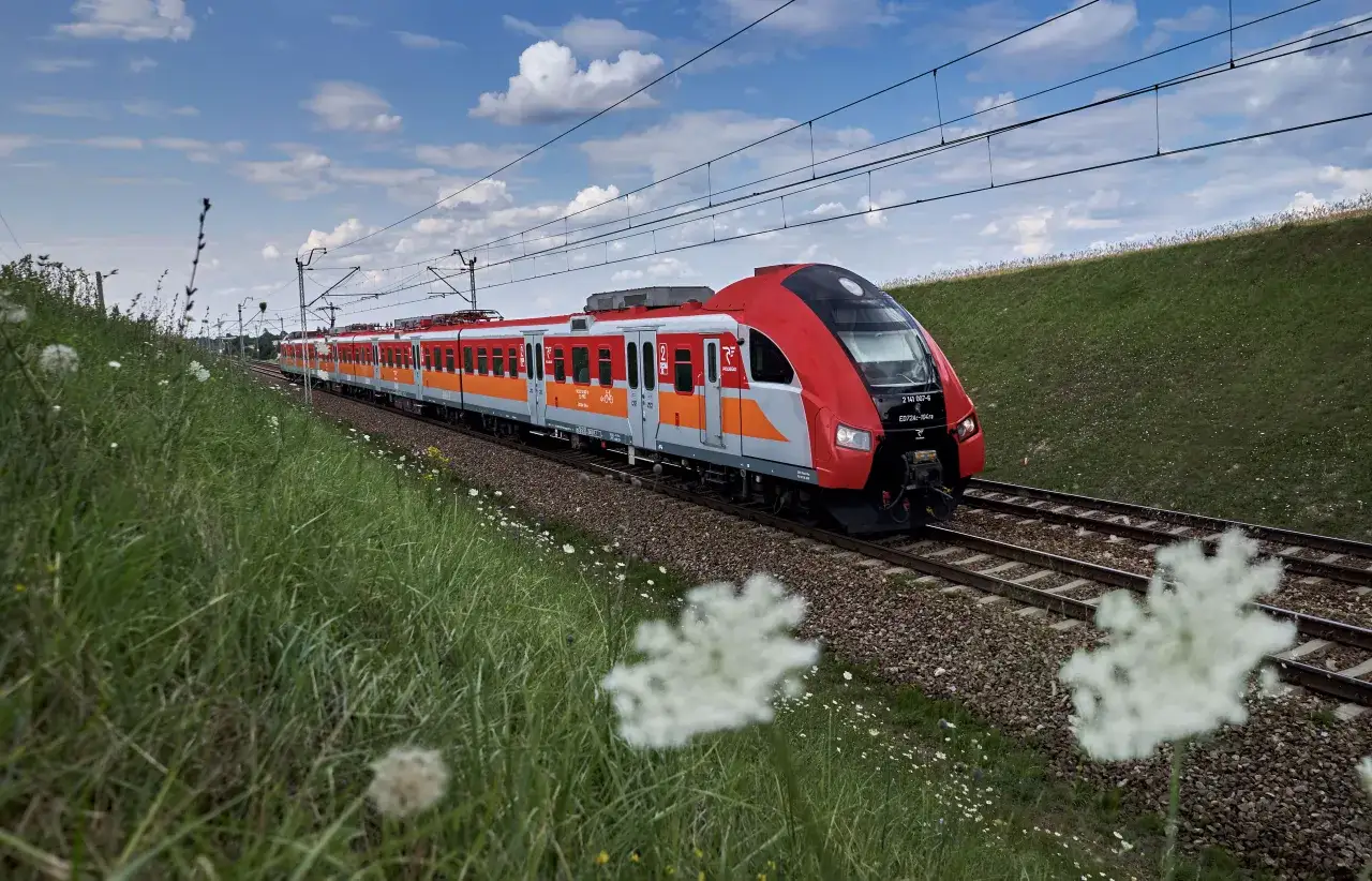 Czerwono-pomarańczowy pociąg mknie po torach wśr&oacute;d zieleni. Idealny środek transportu, gdzie na maj&oacute;wkę pociągiem.