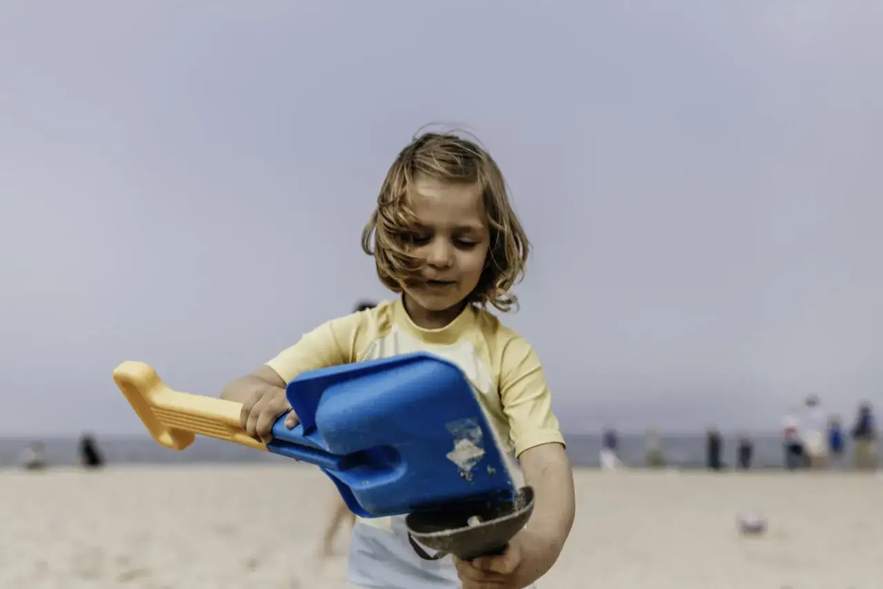 Chłopiec z blond włosami bawi się łopatką na plaży. To idealny przykład, co zabrać na wakacje nad morze z dzieckiem, by zapewnić mu rozrywkę.