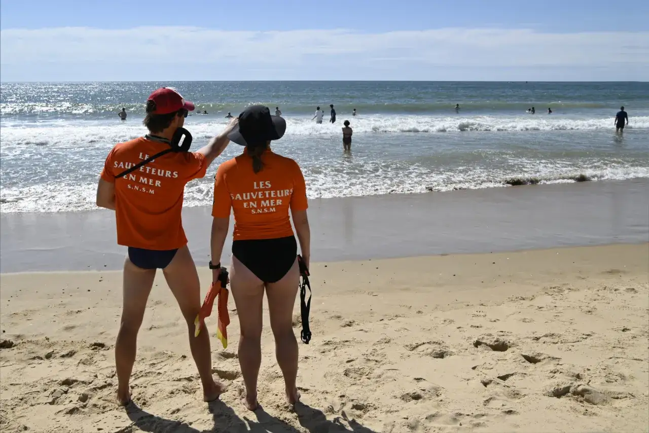 Deux sauveteurs surveillent la **ligne d'eau plage**. L'un porte une casquette rouge, l'autre un chapeau noir. Des baigneurs profitent de la mer.