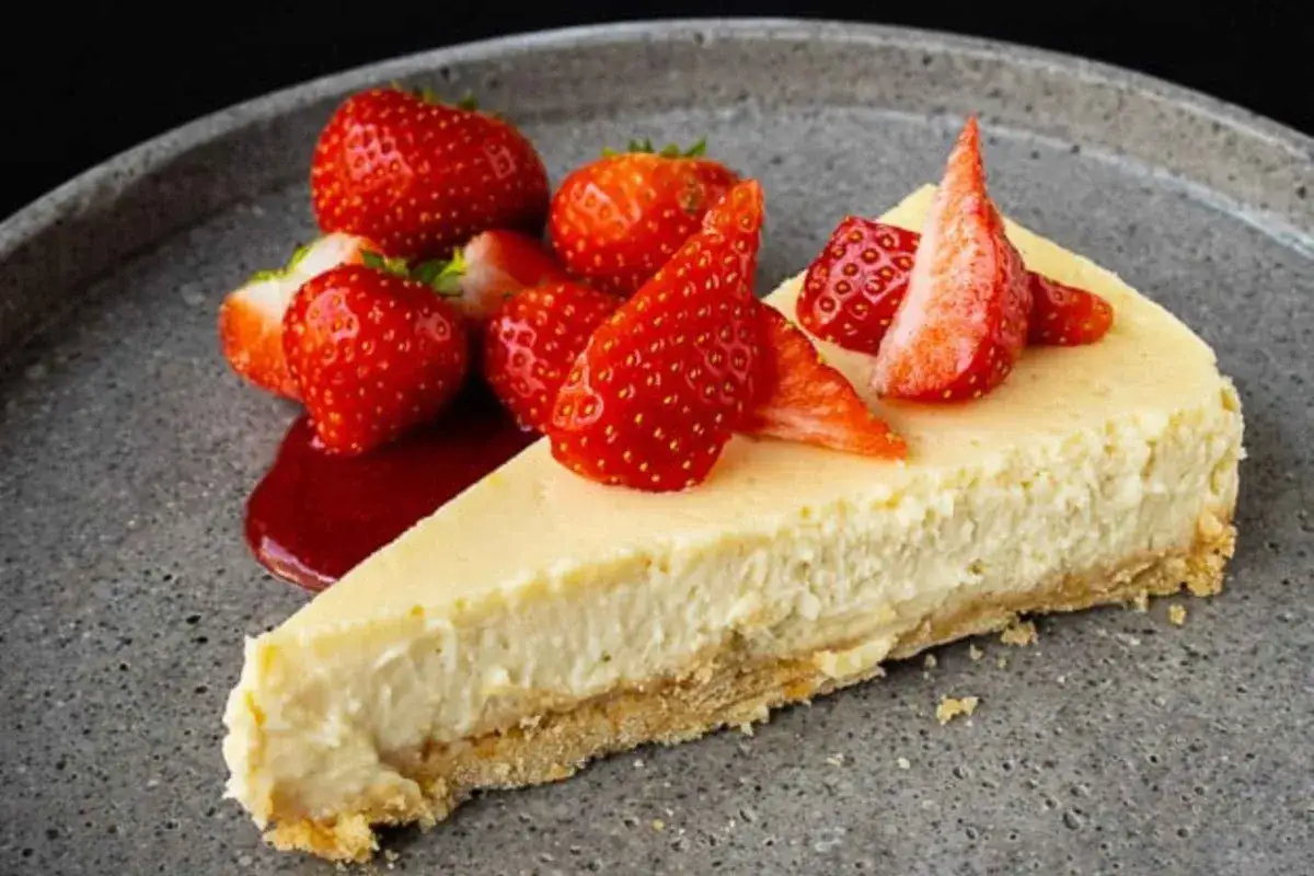 Ein cremiger Quarkkuchen ohne Boden, garniert mit frischen Erdbeeren und Erdbeersauce auf einem grauen Teller.
