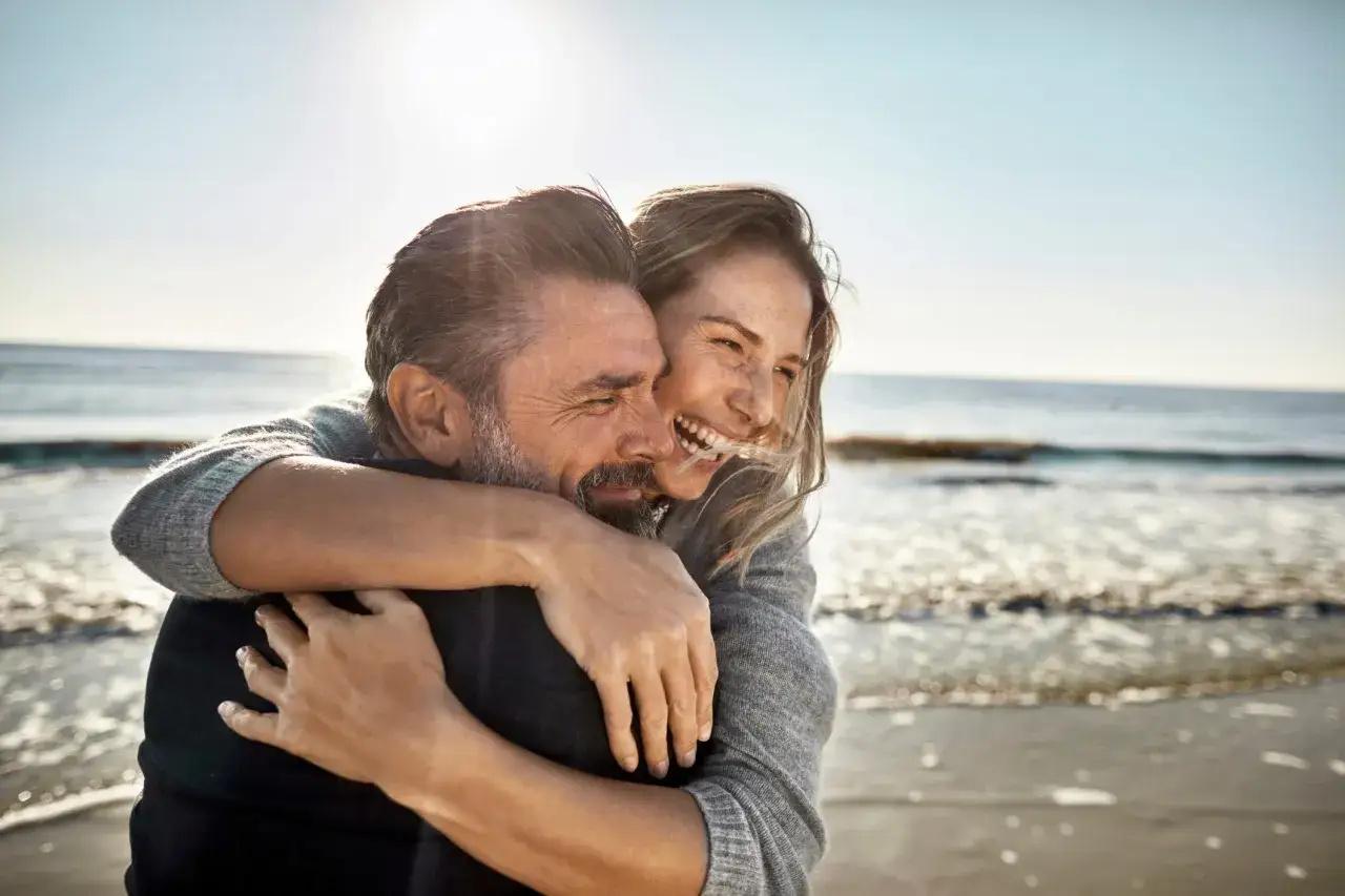 Pareja riendo y abraz&aacute;ndose en la playa. Ella le abraza por detr&aacute;s, &eacute;l sonr&iacute;e. Un ejemplo de como amar.