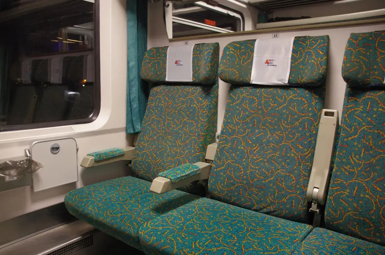 Wnętrze wagonu z przedziałami intercity 2 klasa. Siedzenia w turkusowej tapicerce z pomarańczowym wzorem.