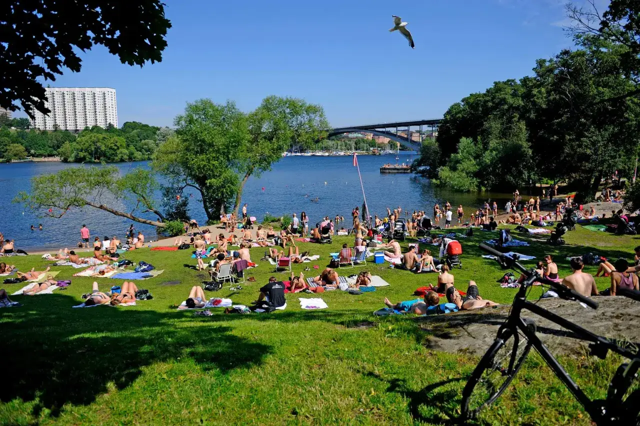 Folk njuter av solen p&aring; en gr&auml;smatta vid en sj&ouml;, en av m&aring;nga popul&auml;ra badplatser i Stockholm.