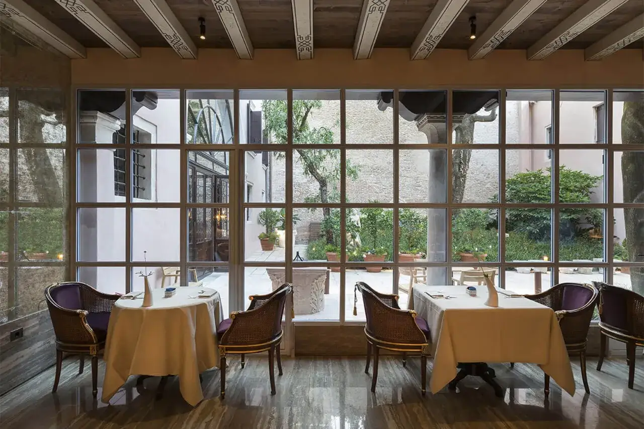 Tavoli apparecchiati in un elegante ristorante stellato Veneto, con vista su un cortile interno con alberi e piante in vaso.