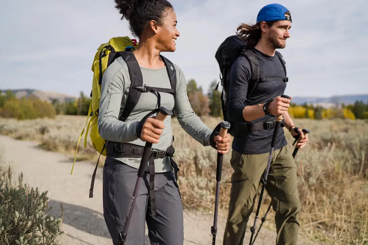Kobieta w stroju w g&oacute;ry damski, z plecakiem i kijkami trekkingowymi, idzie ścieżką wśr&oacute;d suchych traw.
