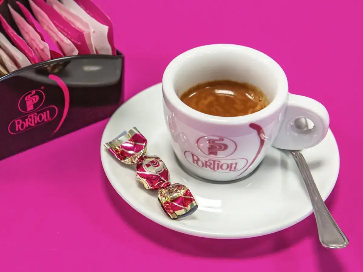 Un'elegante tazzina di caff&eacute; italiano, accompagnata da dolcetti incartati e bustine di zucchero, su un vivace sfondo fucsia.