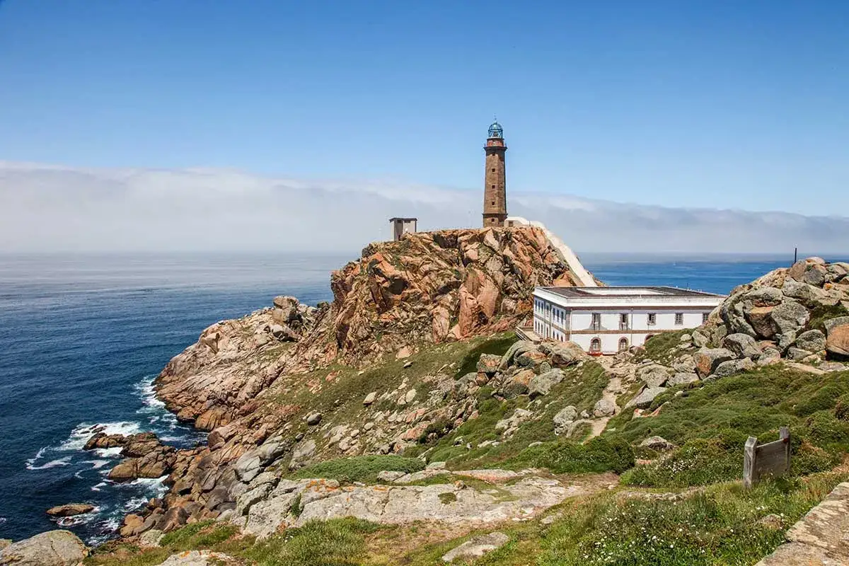 Un faro de piedra se alza sobre un acantilado rocoso junto al mar. Una casa blanca y baja se encuentra a su lado. Ideal para una ruta dos faros en coche.