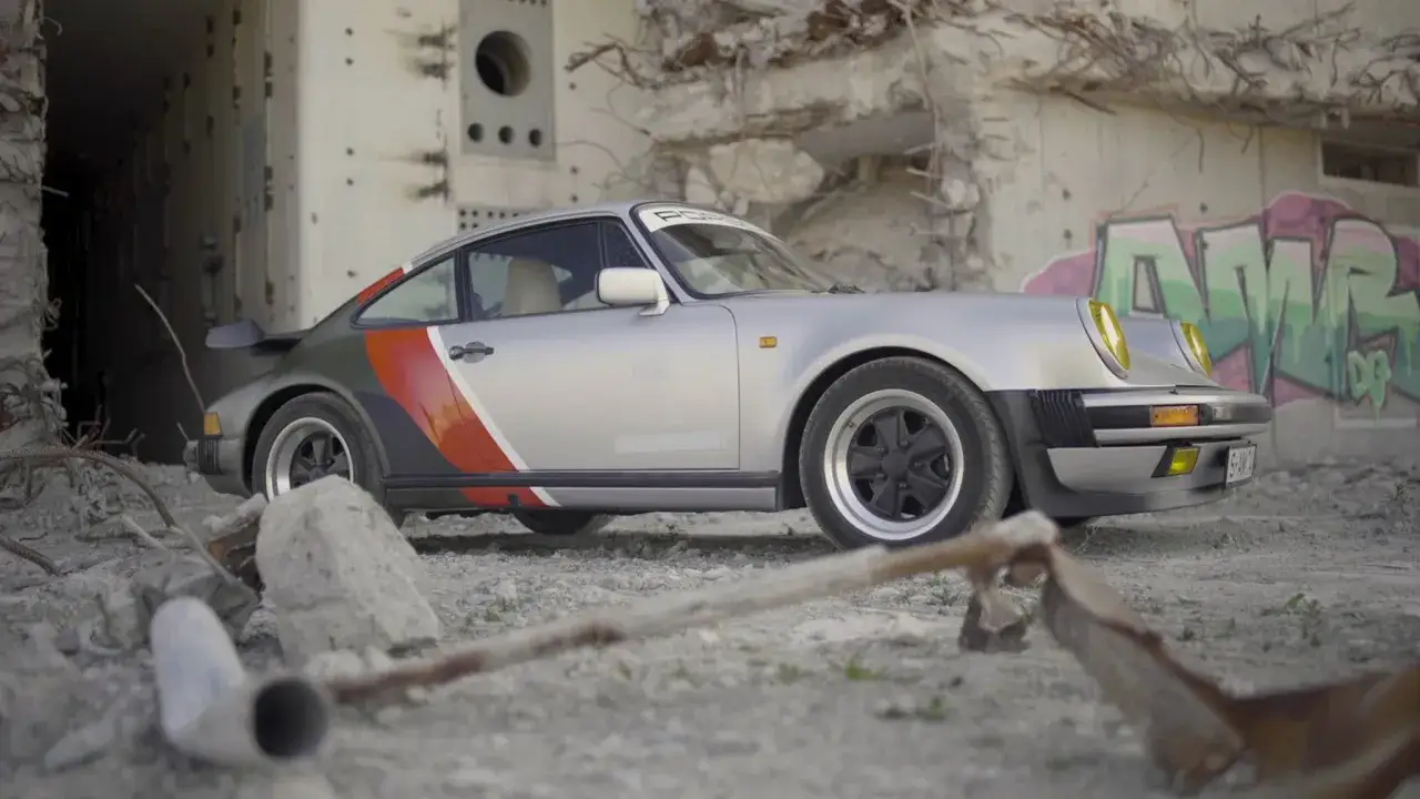 Srebrny Porsche 911 z kolorowymi pasami, stojący wśród ruin, niczym maszyna z przyszłości w świecie cyberpunk.