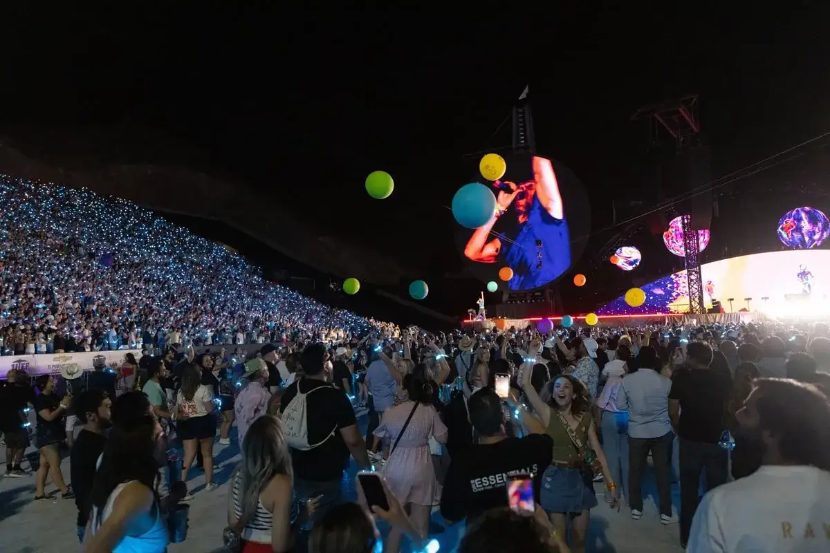 Tłum na koncercie Coldplay, rozświetlony tysiącami świateł, tańczy pod gigantycznym ekranem. Kolorowe kule unoszą się nad sceną.